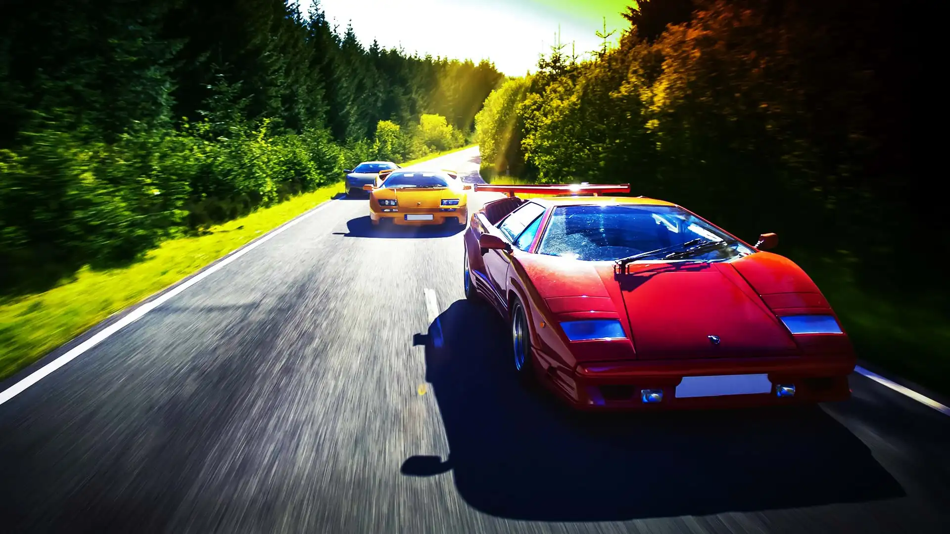 A colorful Lamborghini convoy: Countach 25th Anniversary, Diablo 6.0, and Murciélago, races along a scenic road on a bright day.